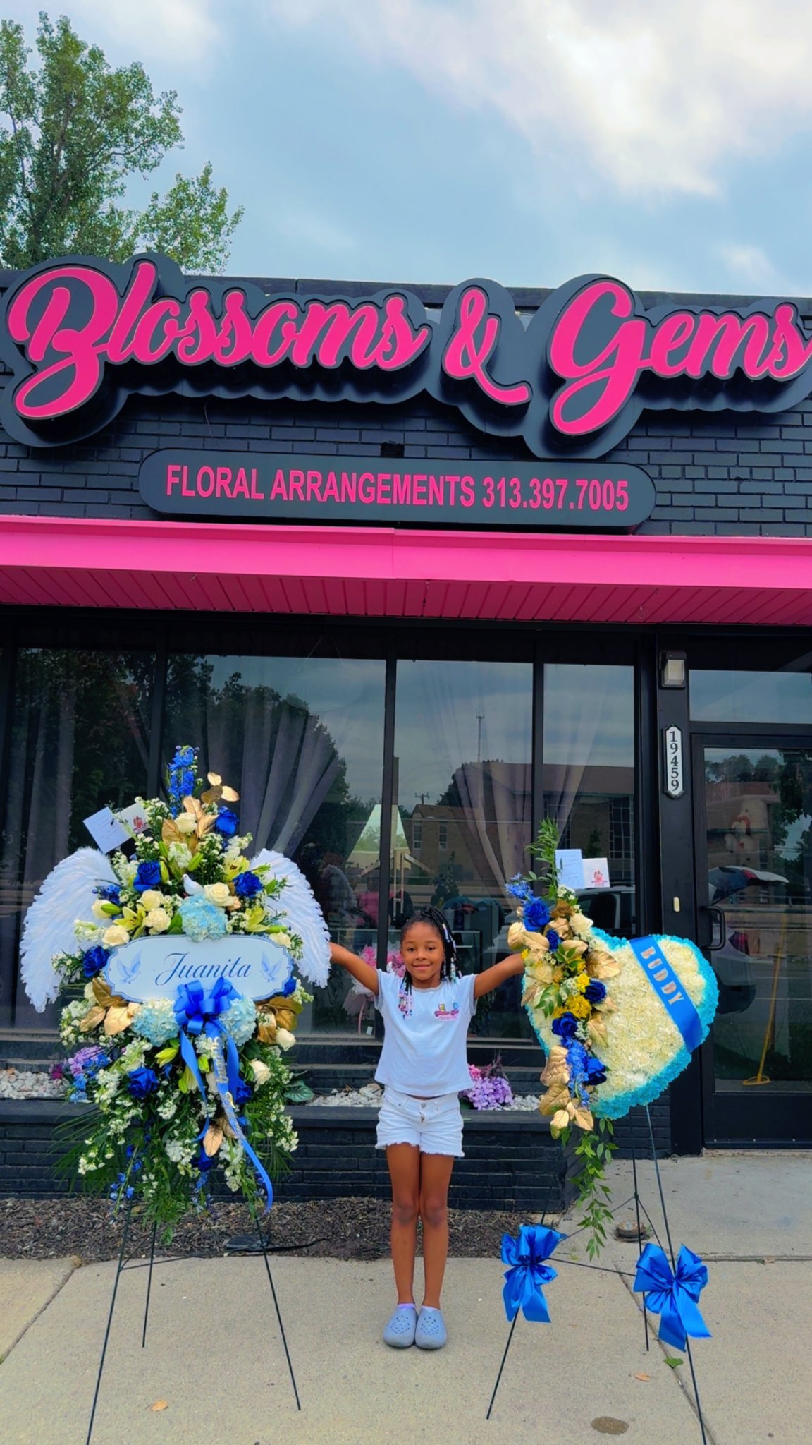 Cali in front of Blossoms & Gems, holding two large floral arrangements under the shop sign